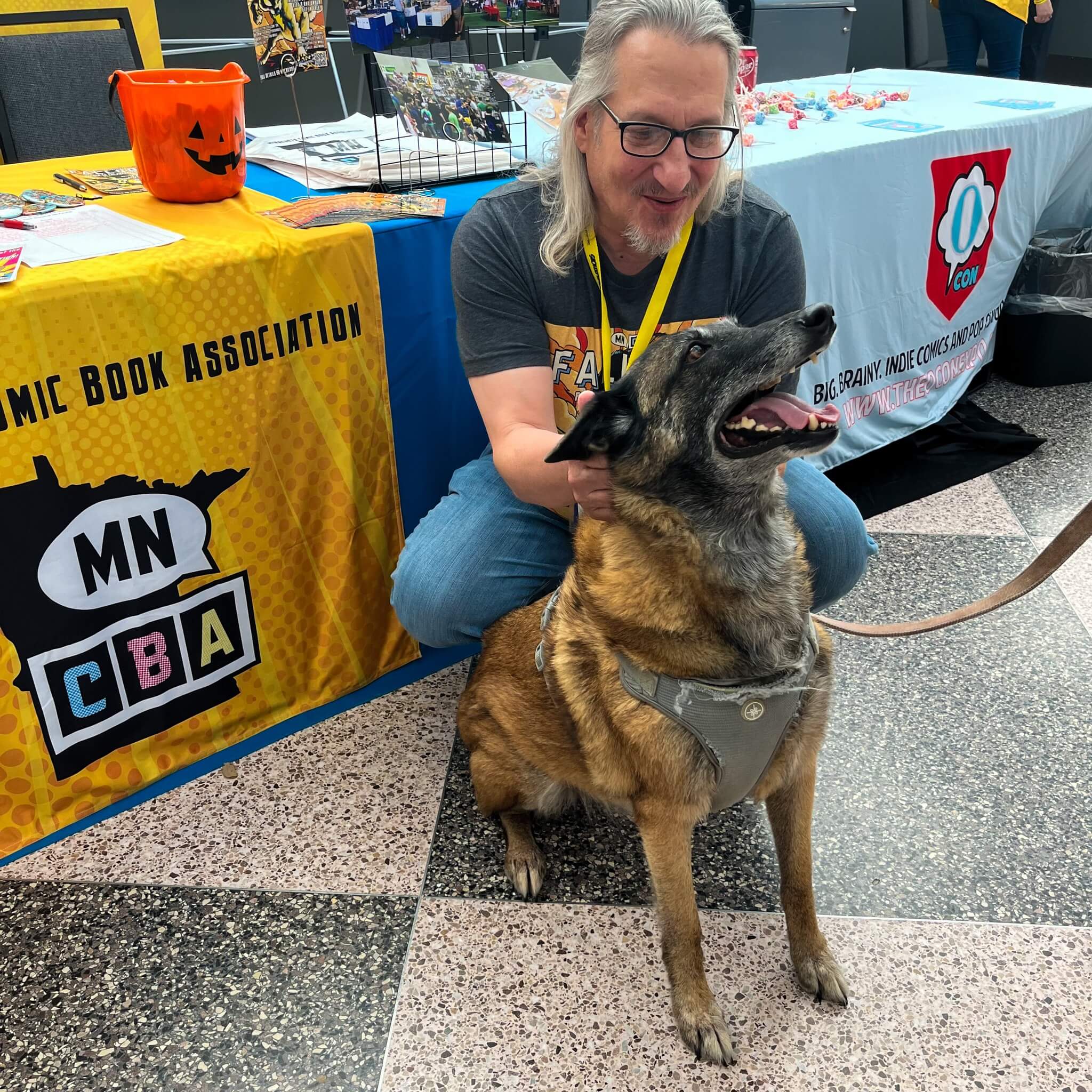 photo of Stephen Henninger kneeling in front of the MNCBA table at an event while petting a dog wearing a harness