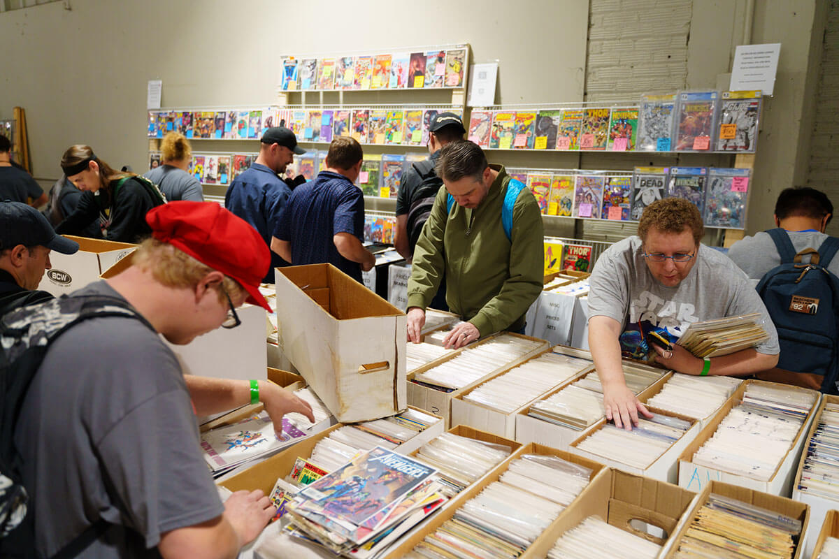 a small crowd of attendees searching for comic books among tables filled with shortboxes